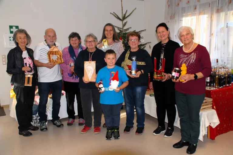 Eine Gruppe von Menschen steht in einem Raum und lächelt für ein Foto. Sie halten verschiedene Gegenstände wie Geschenke, eine Kerze und einen herzförmigen Gegenstand. Hinter ihnen steht ein Weihnachtsbaum.