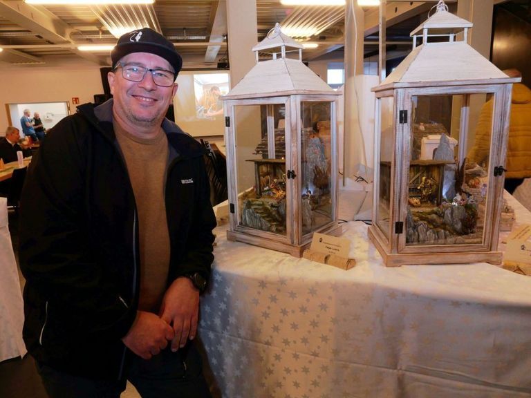 Ein Mann in einer schwarzen Jacke posiert neben zwei Laternen auf einem Tisch und zeigt detaillierte Krippenszenen darin.