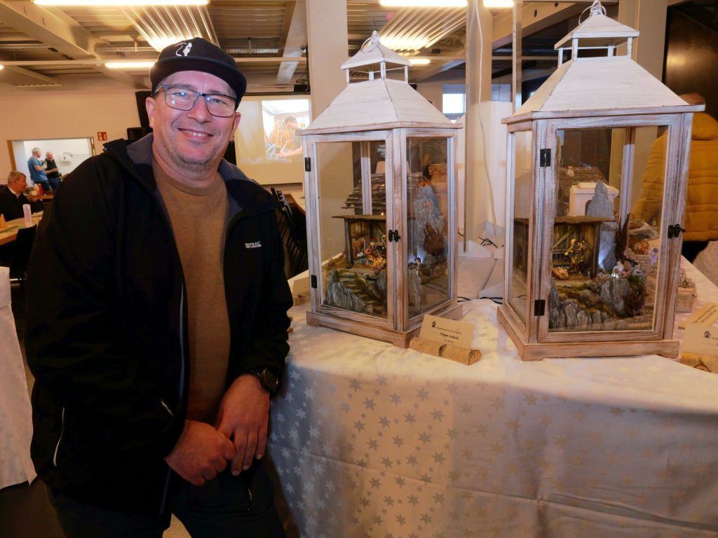Ein Mann in einer schwarzen Jacke posiert neben zwei Laternen auf einem Tisch und zeigt detaillierte Krippenszenen darin.