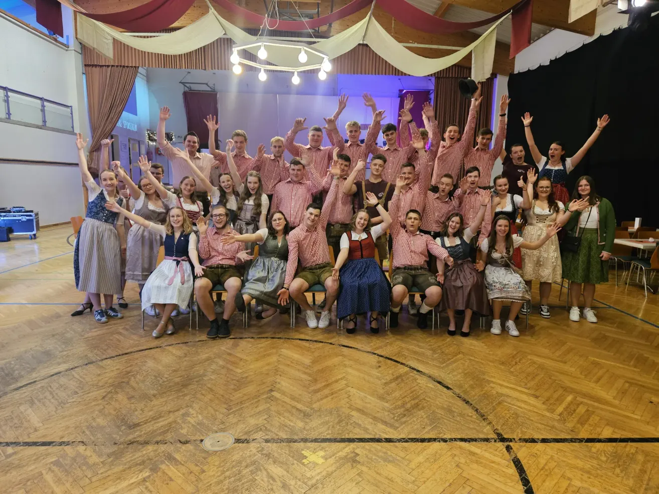 Eine Gruppe junger Erwachsener in traditioneller Kleidung posiert für ein Foto auf einem hölzernen Tanzboden, einige stehend und andere sitzend.