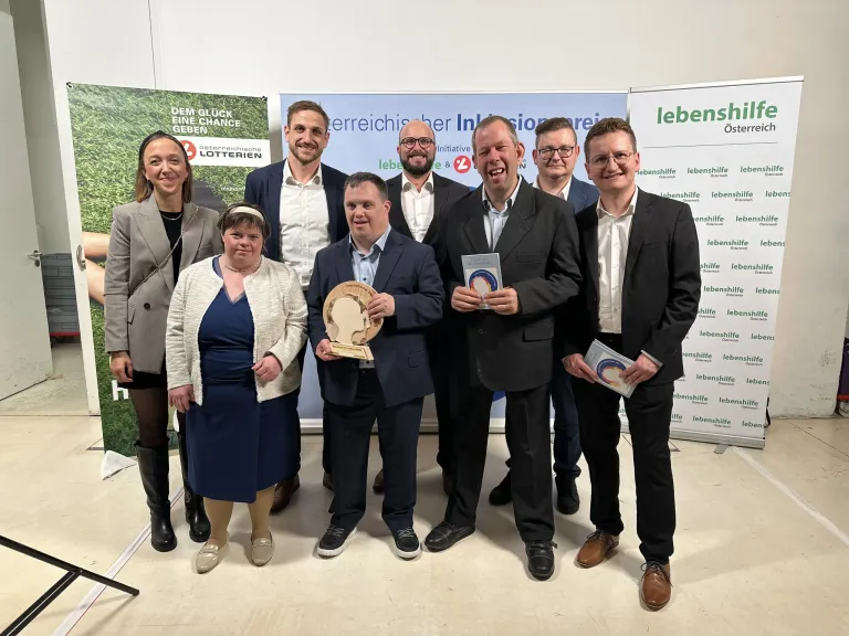 Sieben Personen stehen für ein Foto, zwei davon halten Auszeichnungen. Sie tragen formelle Kleidung. Hinter ihnen befindet sich ein Banner mit Logos und Text. Die Umgebung deutet auf eine Preisverleihung hin.