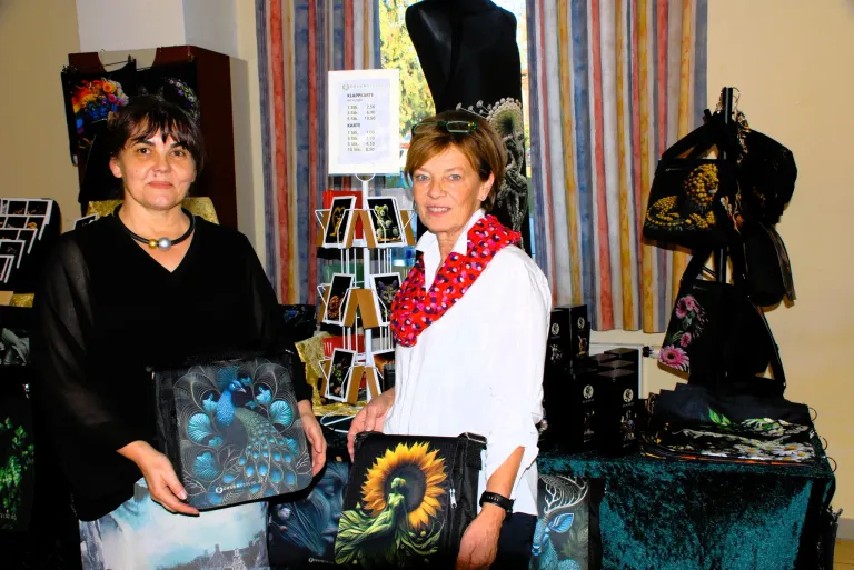 Zwei Frauen stehen vor einem Geschäft, eine hält eine Tasche mit Pfauendesign und die andere eine Tasche mit Sonnenblumen. Dahinter ist eine Ausstellung von Taschen und ein Preisschild ist an einem Ständer befestigt.