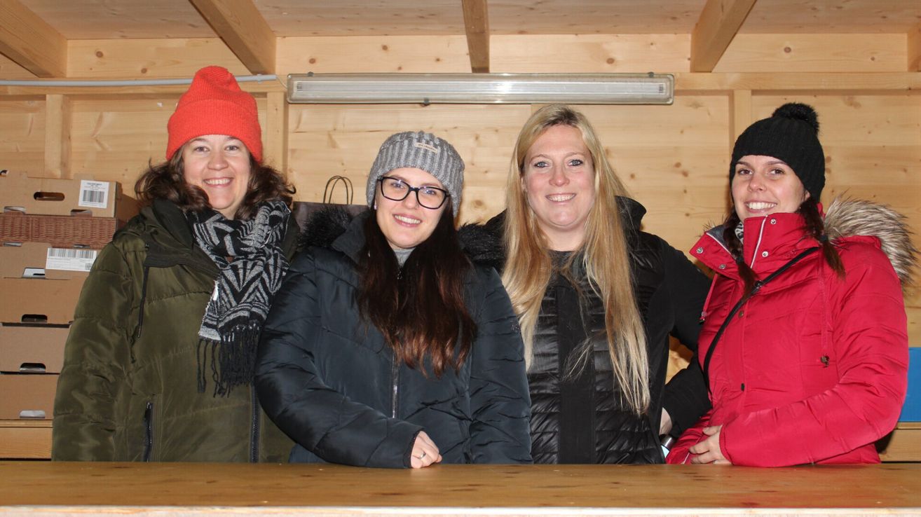 Drei Frauen in Winterkleidung posieren für ein Foto in einer Holzhütte. Die Frau links trägt eine rote Mütze und einen Schal, die mittlere Frau trägt eine graue Mütze und eine Brille und die Frau rechts trägt eine schwarze Jacke.
