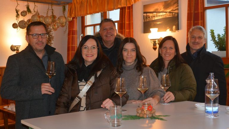 Fünf Erwachsene lächeln und posieren für ein Foto. Sie sitzen um einen Tisch mit Weingläsern und einer Kerze. Die Frau links trägt einen braunen Mantel und eine Brille. Die Wand hat Vorhänge, eine Lampe und ein Foto.