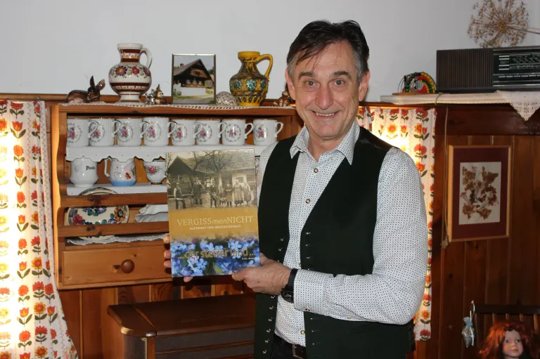 Ein Mann lächelt und hält ein Buch mit dem Titel Vergissmeinnicht in der Hand, vor einem Holzschrank mit verschiedenen Gegenständen darauf.