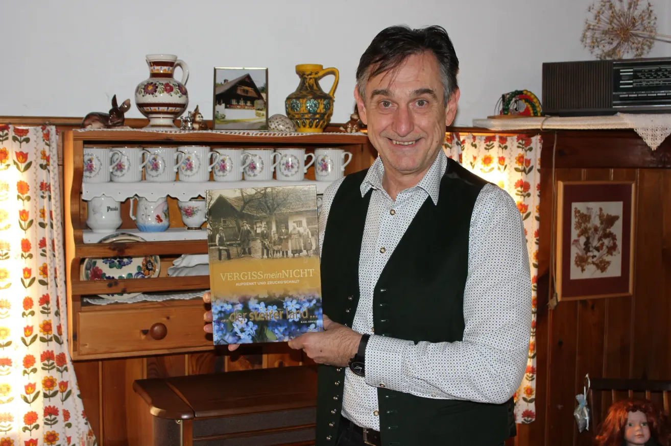Ein Mann lächelt und hält ein Buch mit dem Titel Vergissmeinnicht in der Hand, vor einem Holzschrank mit verschiedenen Gegenständen darauf.