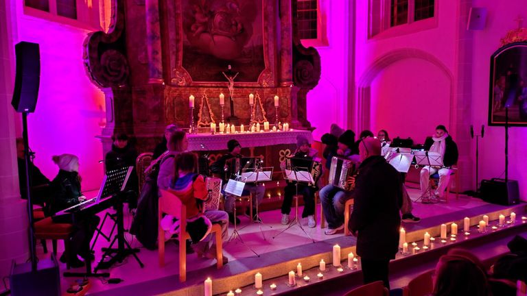 Eine Gruppe von Musikern spielt Akkordeons in einer Kirche mit rosa Beleuchtung. Kerzen sind auf der Bühne und dem Altar angezündet. Die Musiker sind von Zuschauern umgeben.
