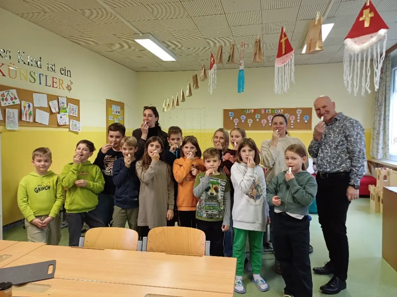 Eine Gruppe von Kindern und Erwachsenen versammelt sich in einem Klassenzimmer. Sie halten Zigaretten und scheinen für ein Foto zu posieren. Hinter ihnen ist der Raum mit verschiedenen Gegenständen dekoriert.