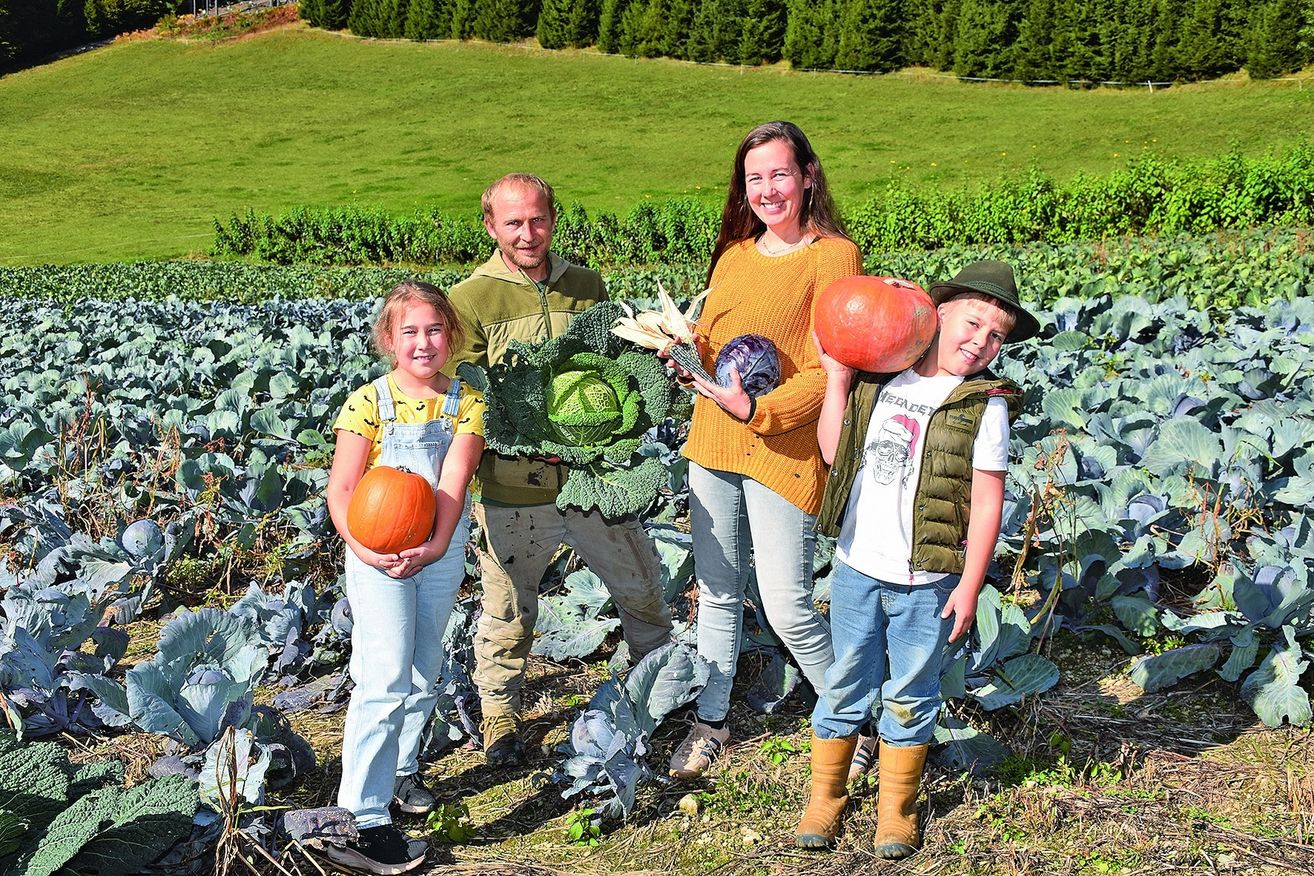 Eine Familie steht in einem Gemüsegarten, lächelt und hält Kürbisse und Kohl. Im Hintergrund ist ein großes grünes Feld und Reihen von Bäumen zu sehen.