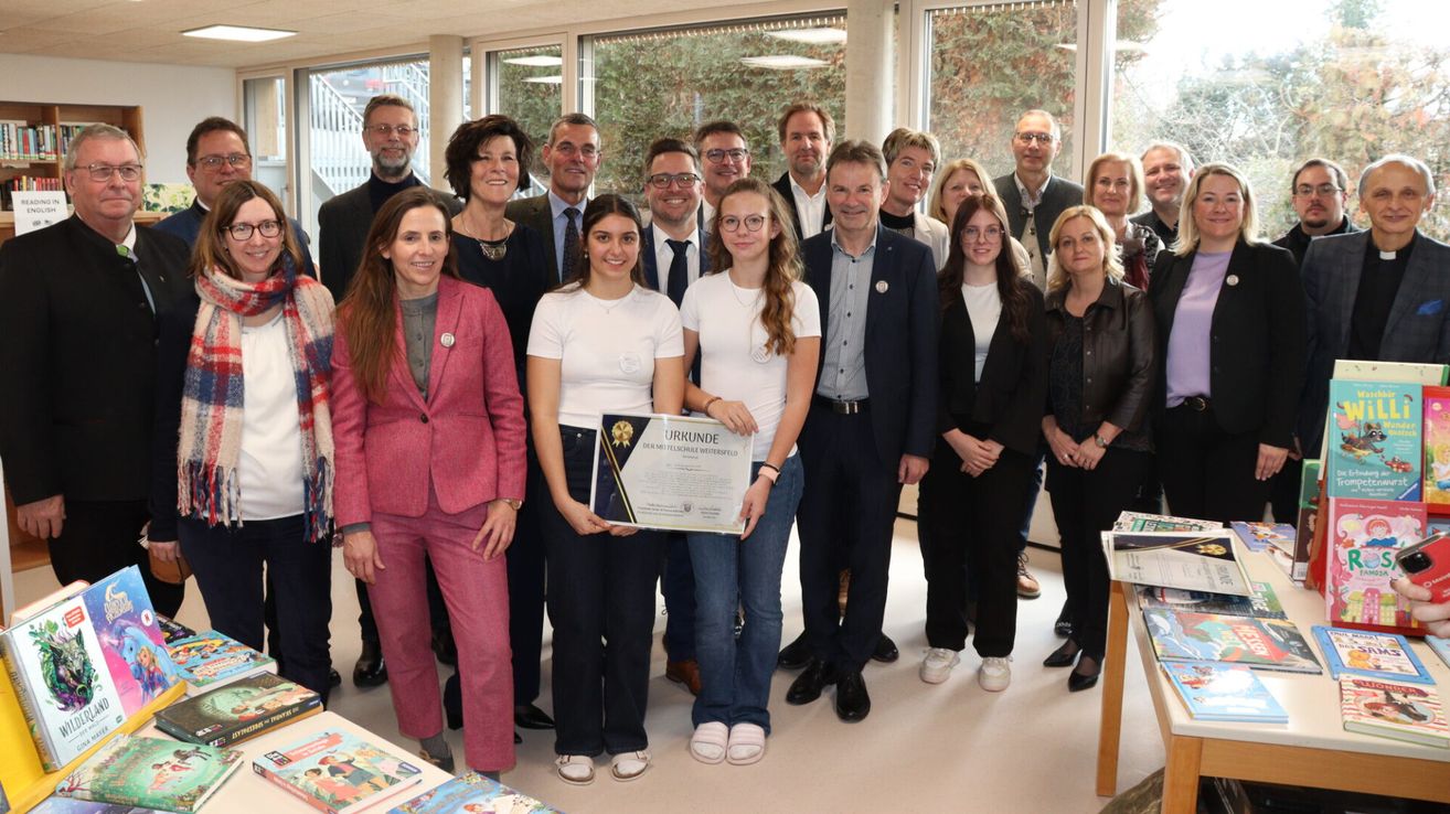 Eine Gruppe von Menschen steht zusammen in einem Innenraum, möglicherweise für ein Gruppenfoto. Zwei Mädchen halten ein Zertifikat, und ein Mann steht vor ihnen. Sie alle lächeln und posieren für die Kamera. Dahinter befinden sich Glasfenster mit Bäumen im Freien.