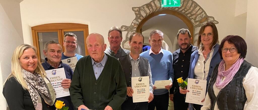 Eine Gruppe von Menschen posiert für ein Foto, lächelt und hält Urkunden in den Händen. Ein älterer Mann sticht heraus, trägt einen grünen Pullover. Eine Frau hält Blumen. Dahinter rahmt ein steinerner Bogen ein grünes Ausfahrtsschild ein.