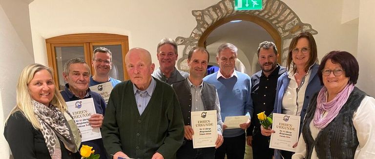 Eine Gruppe von Menschen posiert für ein Foto, lächelt und hält Urkunden in den Händen. Ein älterer Mann sticht heraus, trägt einen grünen Pullover. Eine Frau hält Blumen. Dahinter rahmt ein steinerner Bogen ein grünes Ausfahrtsschild ein.