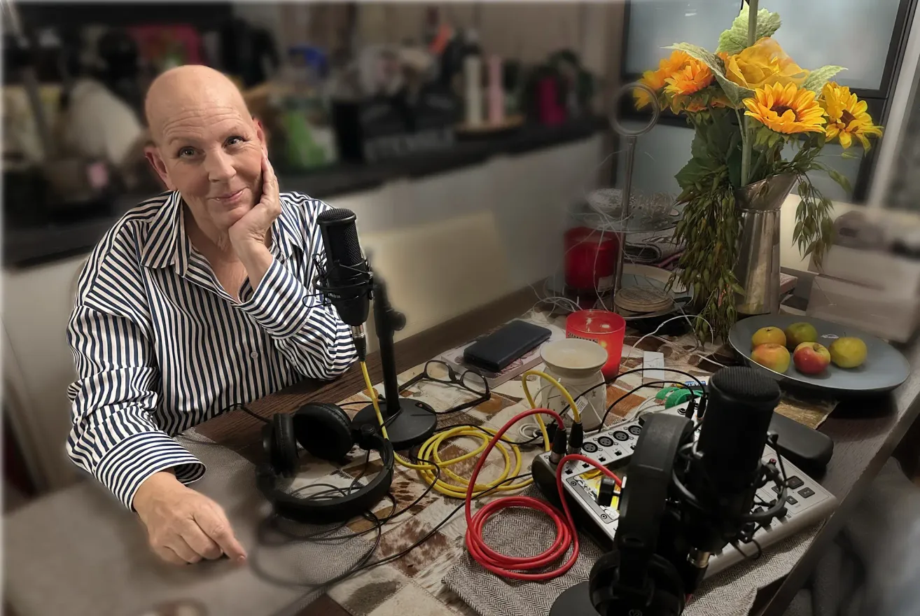 Eine Person mit kahlem Kopf sitzt an einem Schreibtisch mit Mikrofon, Kopfhörern und einem verkabelten Gerät. Der Tisch hat eine karierte Tischdecke, einen Teller, eine Blumenvase und Kerzen. Hinter der Person steht ein Regal mit Flaschen und einer verschwommenen Ansicht des Raums.