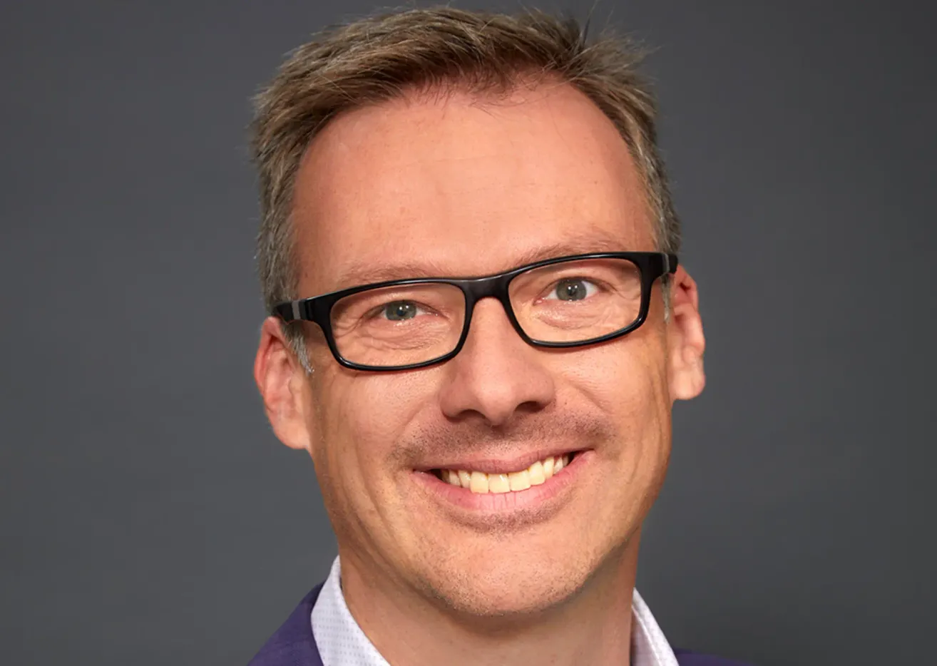 Ein lächelnder Mann mit kurzen Haaren und einer Brille schaut in die Kamera. Er trägt ein helles Hemd unter einer dunklen Jacke.