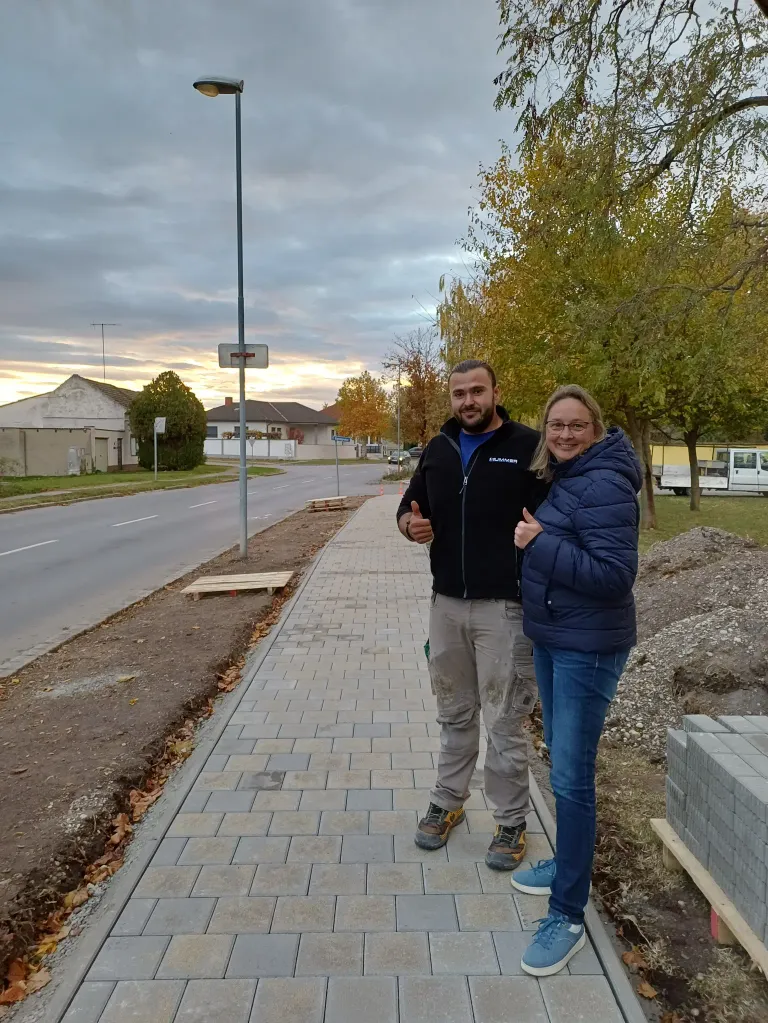 Ein Mann und eine Frau stehen auf einem neu verlegten Ziegelpflaster und lächeln für die Kamera. Sie tragen Winterkleidung, und der Mann gibt einen Daumen hoch. Hinter ihnen befinden sich Bäume, eine Straße und ein Gebäude.