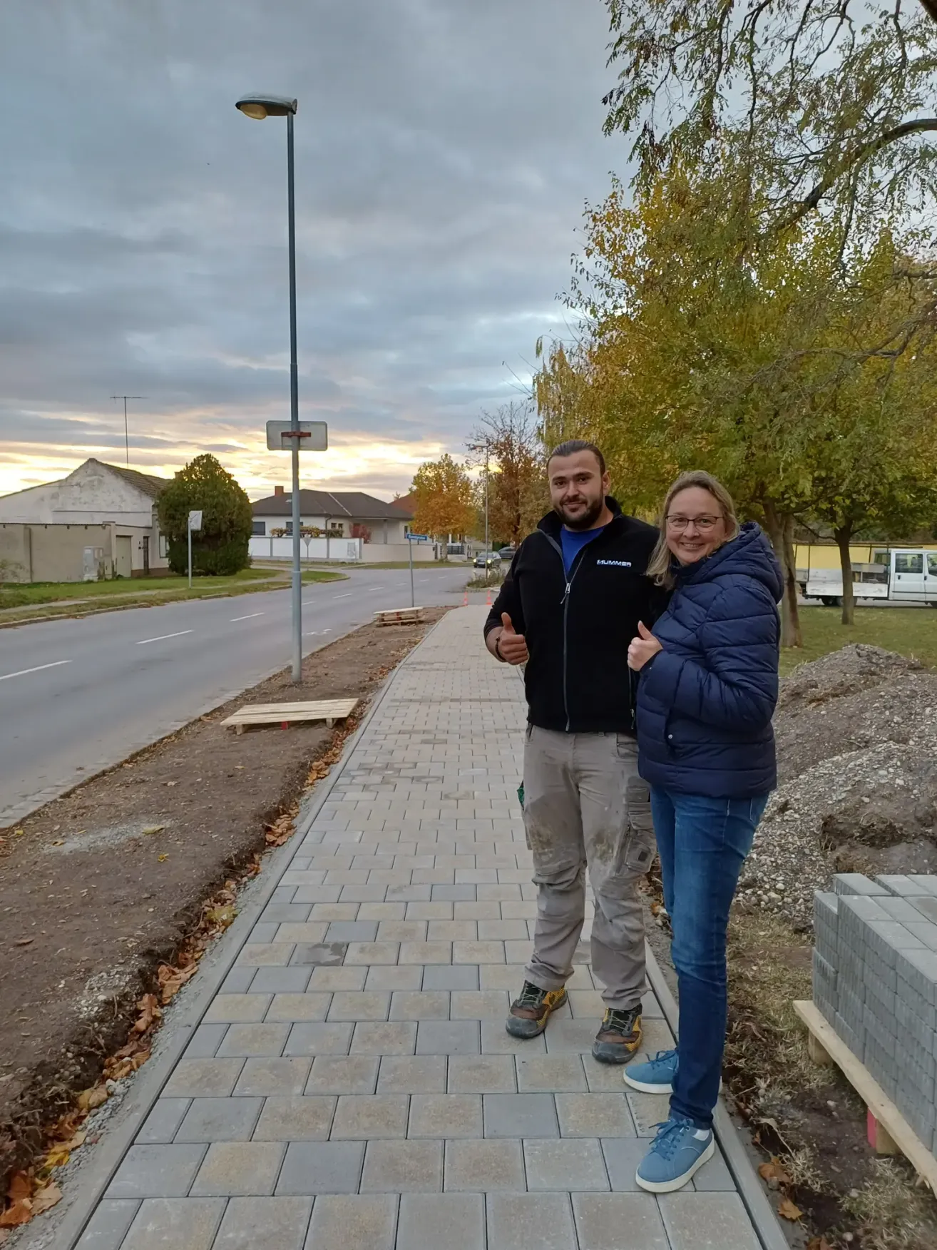 Ein Mann und eine Frau stehen auf einem neu verlegten Ziegelpflaster und lächeln für die Kamera. Sie tragen Winterkleidung, und der Mann gibt einen Daumen hoch. Hinter ihnen befinden sich Bäume, eine Straße und ein Gebäude.
