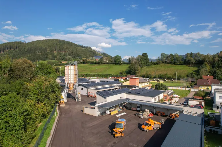 Luftaufnahme eines Fabrikkomplexes mit einem großen Silo, Solarpanelen und Baufahrzeugen in ländlicher Umgebung. Berge und ein klarer Himmel im Hintergrund.