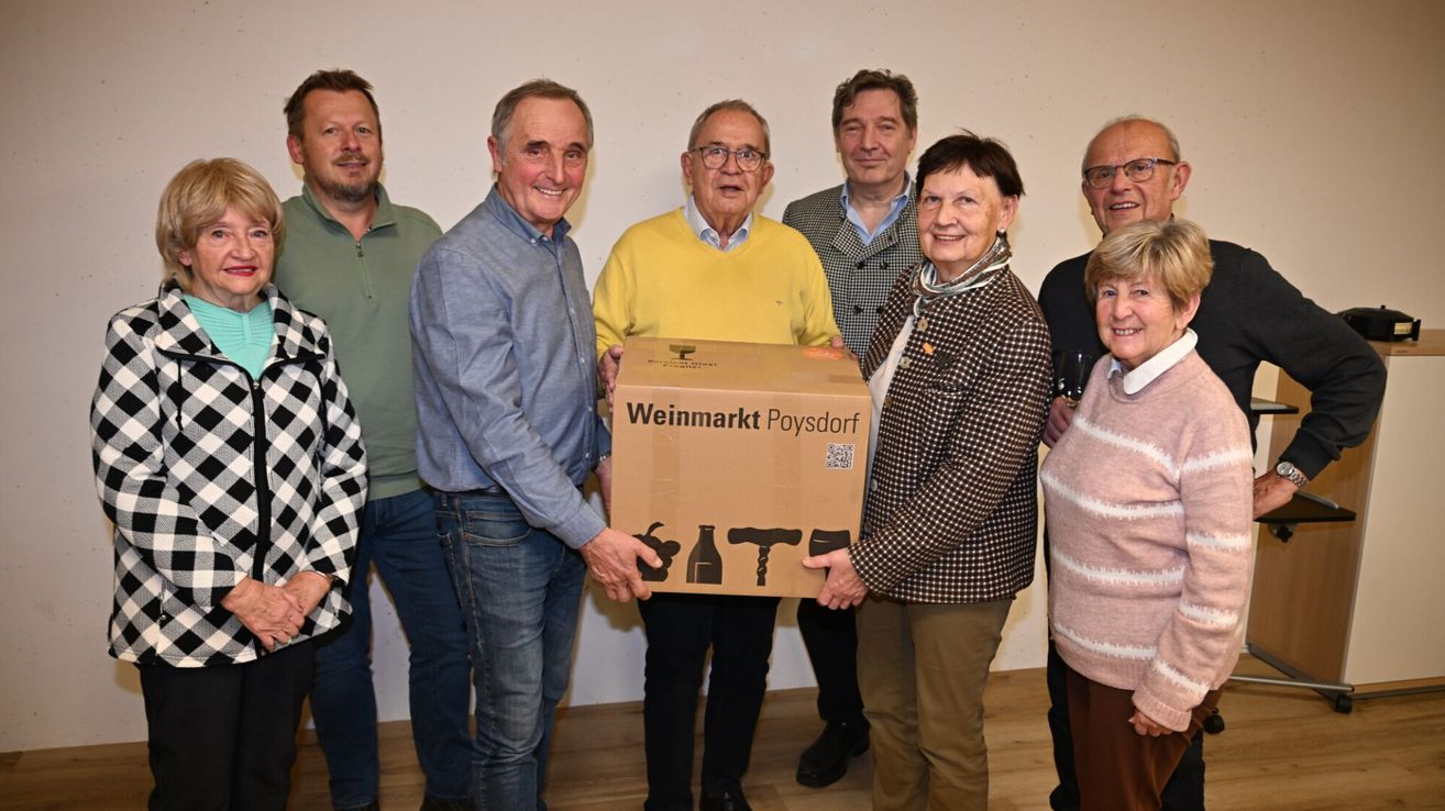 Eine Gruppe von Menschen steht lächelnd zusammen, wobei zwei Personen eine mit Weinmarkt Poysdorf beschriftete Box präsentieren. Die Box enthält Weinflaschen und einen Korkenzieher.