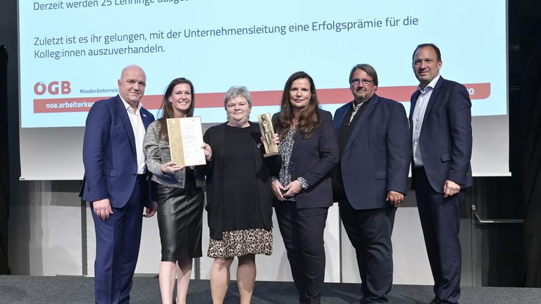 Fünf Personen stehen zusammen und halten Auszeichnungen in der Hand, lächeln für ein Foto. Ein großer Bildschirm hinter ihnen zeigt einen deutschen Text.