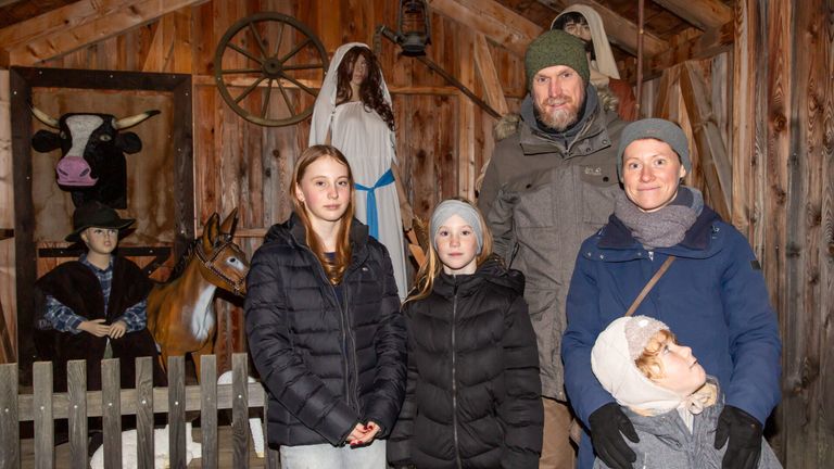 Eine Familie posiert für ein Foto in einer rustikalen Umgebung mit einem Holzrad und Krippenfiguren im Hintergrund. Zwei junge Mädchen und zwei Erwachsene stehen zusammen.