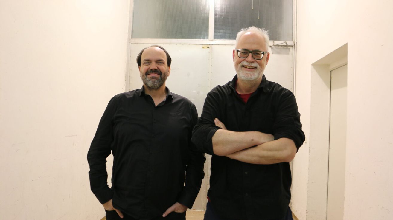 Zwei Männer in schwarzen Hemden stehen nah beieinander, einer mit den Händen in den Taschen, der andere mit verschränkten Armen, lächelnd, vor einer weißen Wand.