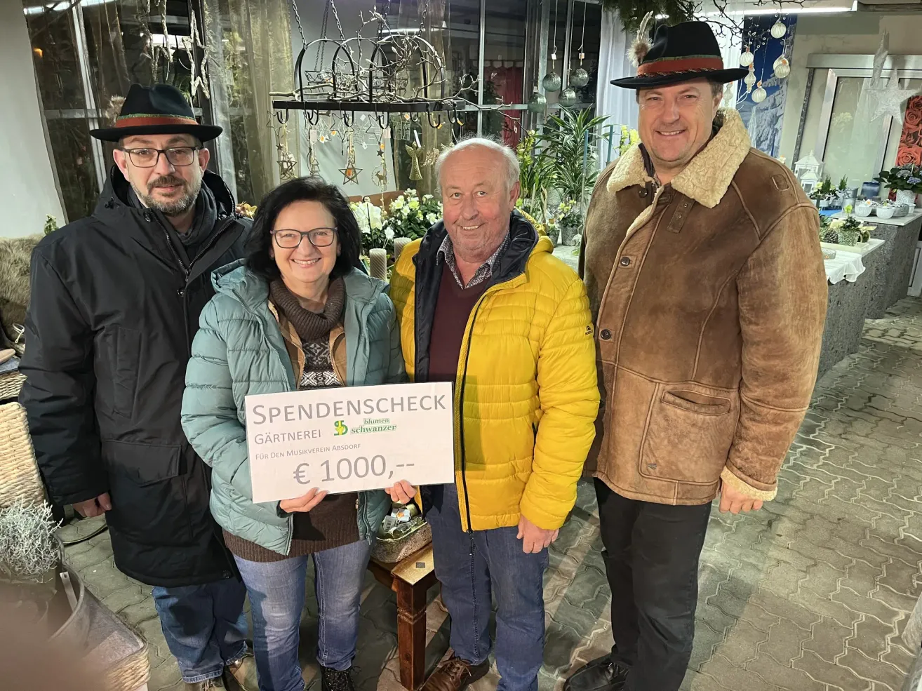 Vier Personen stehen in einem Raum mit einem von einer Frau gehaltenen Banner. Das Banner trägt den Aufdruck 'Spendenscheck für den Musikverein Absdopf' mit einem Scheck über 1000 Euro. Hinter ihr lächelt ein Mann in einer gelben Jacke. Der Mann auf ihrer linken Seite trägt einen schwarzen Hut und eine Brille.