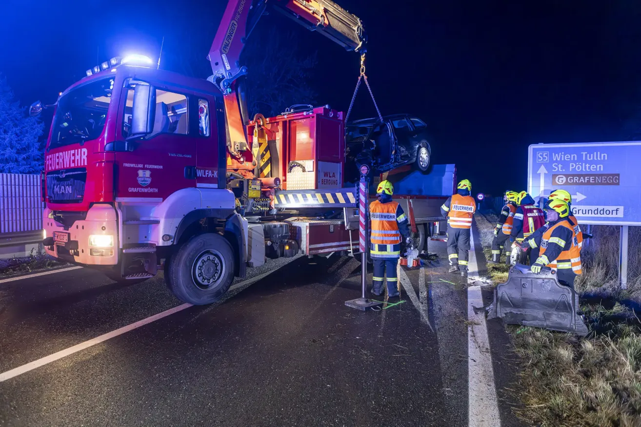 Ein Feuerwehrwagen mit einem Kran entfernt ein beschädigtes Auto von der Straße bei Nacht. Feuerwehrleute stehen um das Fahrzeug herum, zwei von ihnen tragen Helme.