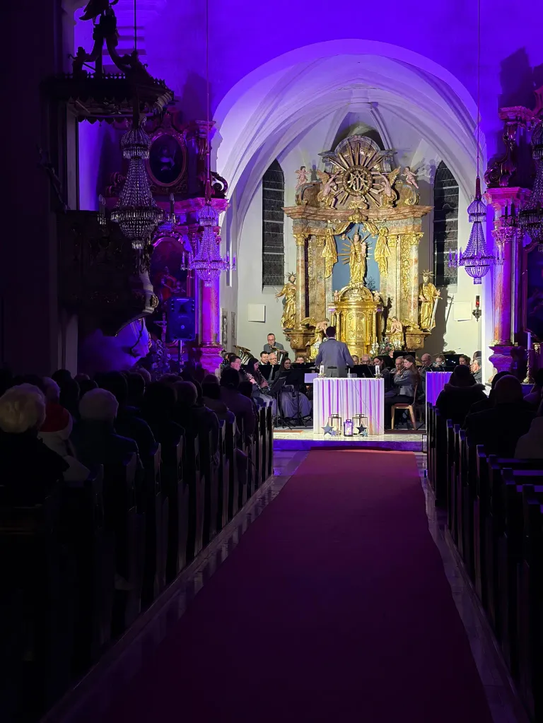 Ein Orchester tritt in einer Kirche auf, das Publikum sitzt auf Kirchenbänken. Die Bühne ist mit einem roten Teppich dekoriert und von goldenen Statuen flankiert.