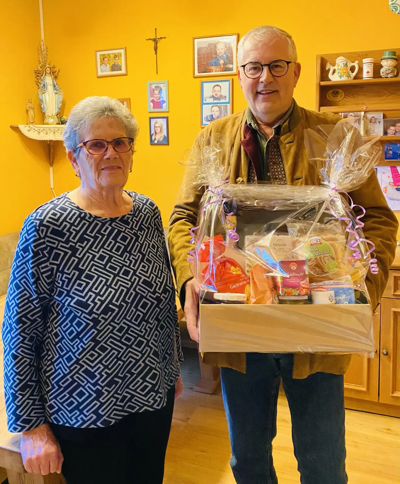 Ein älteres Paar steht in einem Zimmer mit gelber Wand und hält eine Geschenkbox. Die Frau trägt eine Brille und ein gemustertes Hemd. Dahinter befinden sich ein Regal mit Familienfotos und eine Statue.