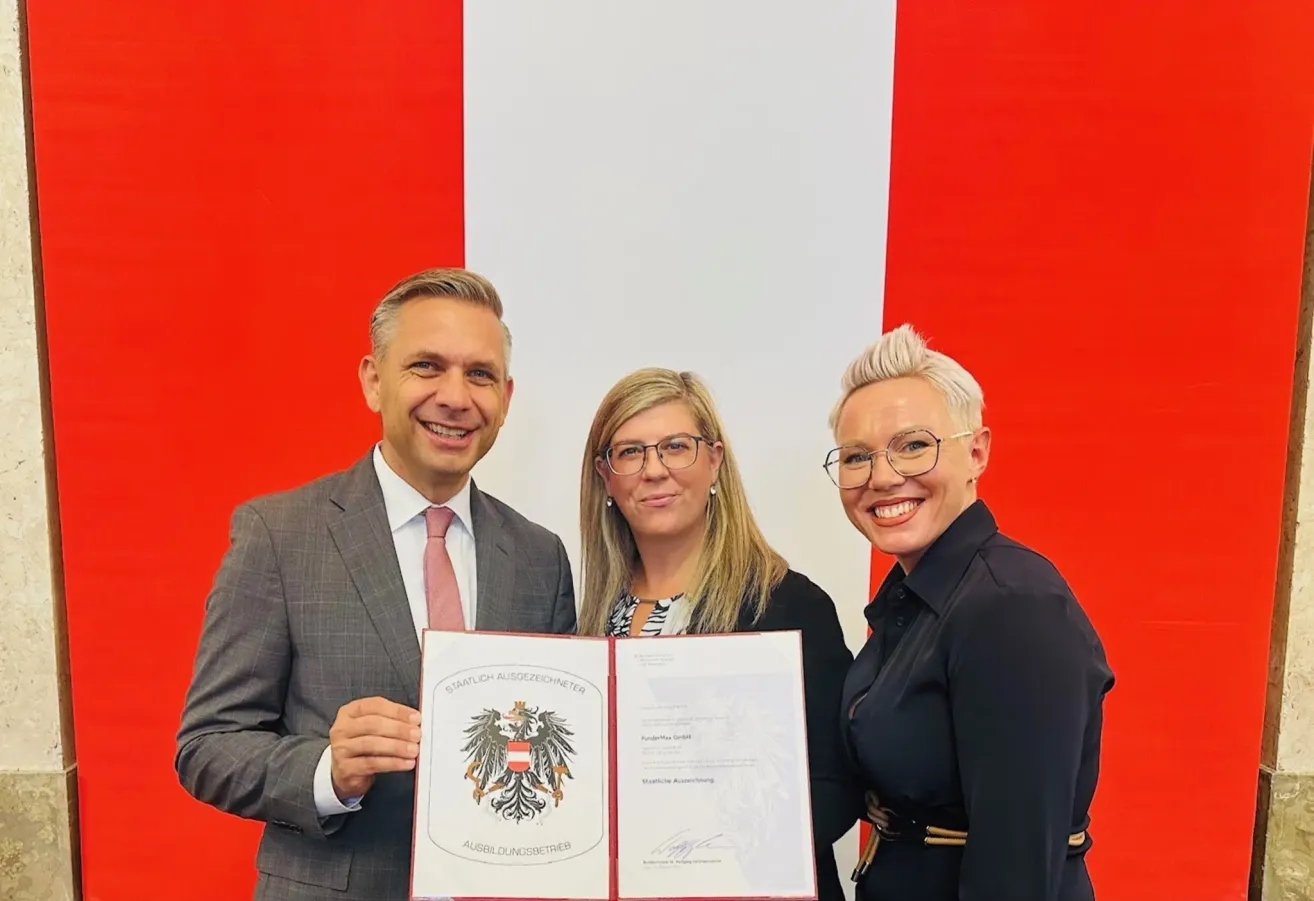 Drei Personen, zwei Frauen und ein Mann, stehen gemeinsam und halten ein Zertifikat. Der Mann trägt einen Anzug und eine Krawatte, und die Frauen tragen eine Brille. Sie alle lächeln. Hinter ihnen ist ein Hintergrund mit der österreichischen Flagge.
