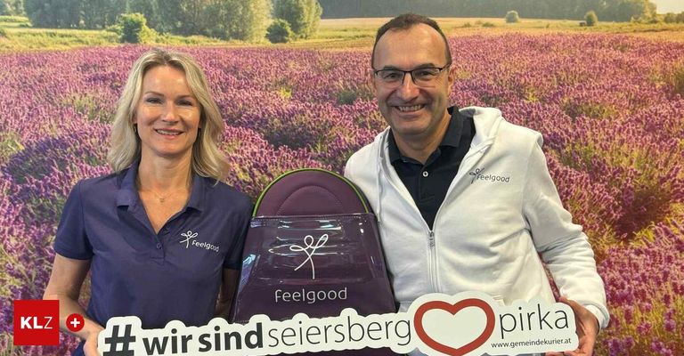 Ein Mann und eine Frau stehen neben einem Lavendelfeld und lächeln. Sie halten eine lila Tasche mit dem Wort 'Feelgood' und einem Logo. Sie tragen passende Polo-Shirts. Ein rotes Herz und Text sind im Vordergrund.