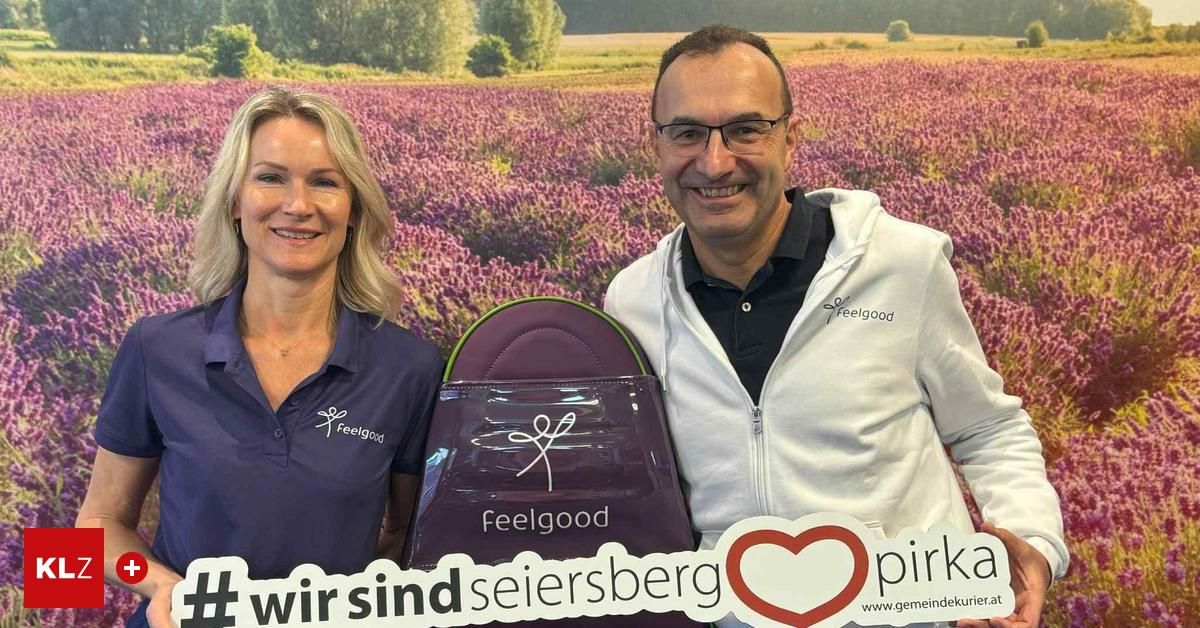 Ein Mann und eine Frau stehen neben einem Lavendelfeld und lächeln. Sie halten eine lila Tasche mit dem Wort 'Feelgood' und einem Logo. Sie tragen passende Polo-Shirts. Ein rotes Herz und Text sind im Vordergrund.