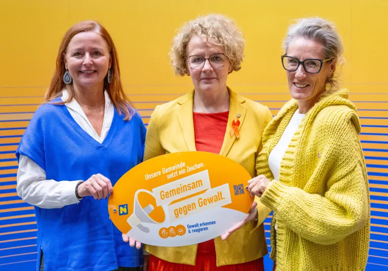 Drei Frauen stehen lächelnd zusammen und halten ein Schild mit der Aufschrift 'Gemeinsam gegen Gewalt'. Die Frau links trägt einen blauen Pullover, die mittlere Frau einen gelben Blazer und die rechte Frau einen gelben Strickpullover. Das Schild hat ein orangefarbenes Band.
