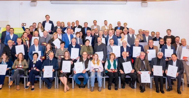 Eine Gruppe von Menschen, die sitzen und stehen, halten Urkunden und Auszeichnungen, wahrscheinlich bei einer Preisverleihung.