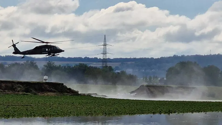 Ein Hubschrauber fliegt tief über einen Fluss und erzeugt eine große Gischt. Bäume und ein Strommast sind im Hintergrund zu sehen.