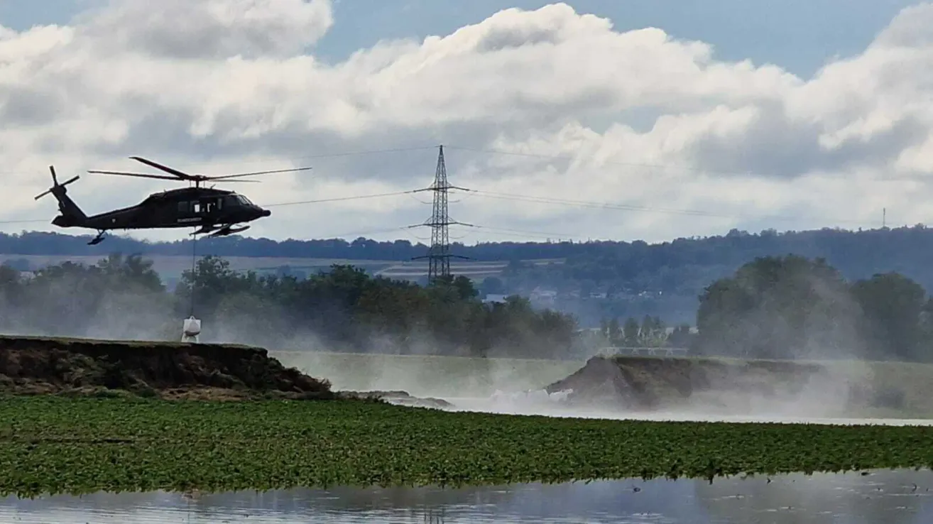 Ein Hubschrauber fliegt tief über einen Fluss und erzeugt eine große Gischt. Bäume und ein Strommast sind im Hintergrund zu sehen.