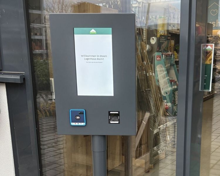 Ein grauer Kiosk mit einem Bildschirm, auf dem 'Willkommen in Ihrem Lagerhaus Markt' steht, und einem blauen Knopf mit Logo steht vor einem Geschäft.