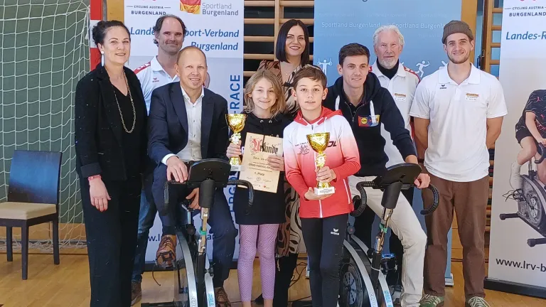 Eine Gruppe von Erwachsenen und Kindern posiert für ein Foto vor einem Hintergrund mit Fahrrad- und Sportthemen. Die Erwachsenen und Kinder halten Pokale und Urkunden.