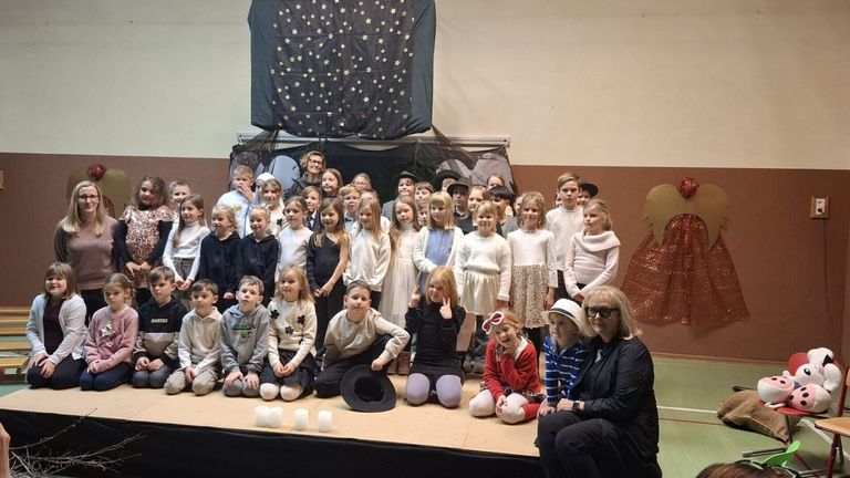 Eine Gruppe von Kindern und zwei Erwachsenen steht auf der Bühne und trägt verschiedene Kostüme. Einige sitzen auf dem Boden, während andere hinter ihnen stehen. Ein sternübersäter Hintergrund und zusätzliche Dekorationen befinden sich hinter der Gruppe.