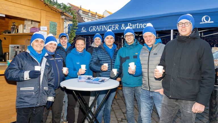 Eine Gruppe von Männern in Winterkleidung, die Tassen halten, steht draußen unter einem blauen Zelt mit dem Namen Grafinger & Partner.