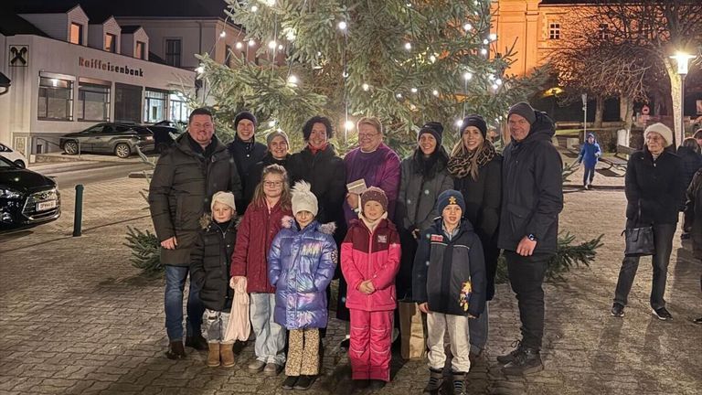 Eine Gruppe von Menschen, die warm gekleidet sind, lächeln und vor einem geschmückten Weihnachtsbaum für ein Foto posieren. Einige halten Geschenke.
