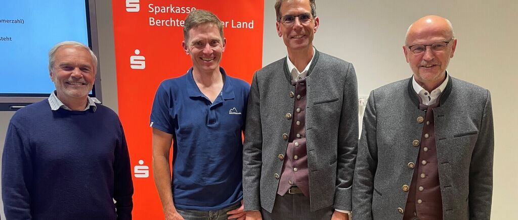 Vier Männer stehen lächelnd vor einem roten Banner mit den Worten 'Sparkasse Berchtesgaden Land'. Der Mann auf der linken Seite trägt ein blaues Hemd, der Mann neben ihm eine graue Jacke. Die beiden Männer auf der rechten Seite tragen eine graue Jacke mit braunen Westen.