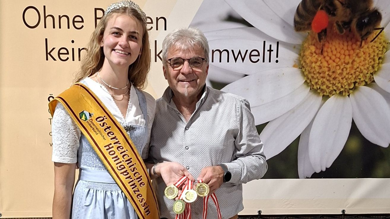 Eine junge Frau mit einer Schärpe, auf der 'Osterreichische Honigprinzessin' steht, und einer Krone lächelt neben einem älteren Mann, der Medaillen hält. Sie stehen vor einer weißen Blume mit den Worten 'Plen kwelt!'.