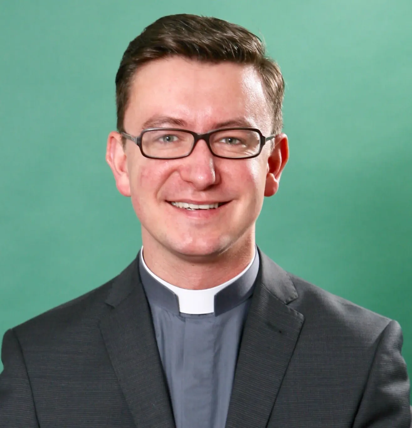 Ein lächelnder Mann im Priesterkragen und mit Brille posiert vor einem grünen Hintergrund.