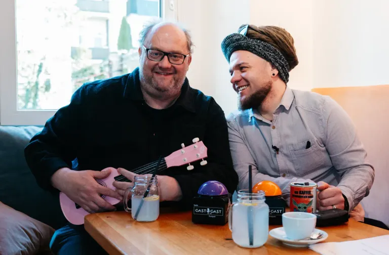 Zwei Männer sitzen an einem Tisch, lächeln und halten Getränke. Einer hält eine rosa Ukulele. Verschiedene Gläser und Tassen stehen auf dem Tisch.
