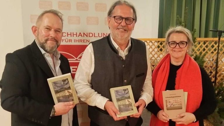 Drei Menschen stehen lächelnd und halten Bücher, möglicherweise bei einer Veranstaltung. Der Mann auf der linken Seite trägt ein gestreiftes Hemd und eine Brille. Der Mann in der Mitte trägt eine Weste und eine Brille, und die Frau auf der rechten Seite trägt einen roten Schal und eine Brille. Hinter ihnen ist ein roter Hintergrund.