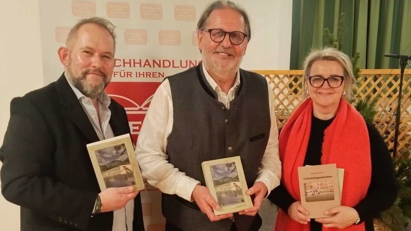 Drei Menschen stehen lächelnd und halten Bücher, möglicherweise bei einer Veranstaltung. Der Mann auf der linken Seite trägt ein gestreiftes Hemd und eine Brille. Der Mann in der Mitte trägt eine Weste und eine Brille, und die Frau auf der rechten Seite trägt einen roten Schal und eine Brille. Hinter ihnen ist ein roter Hintergrund.