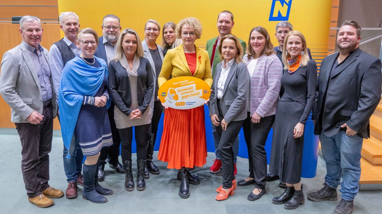 Eine Gruppe von Erwachsenen steht in einem Kreis vor einer gelben Wand. Eine Frau in der Mitte hält ein gelbes Schild. Einige von ihnen tragen eine Brille und einige tragen rote Bänder.