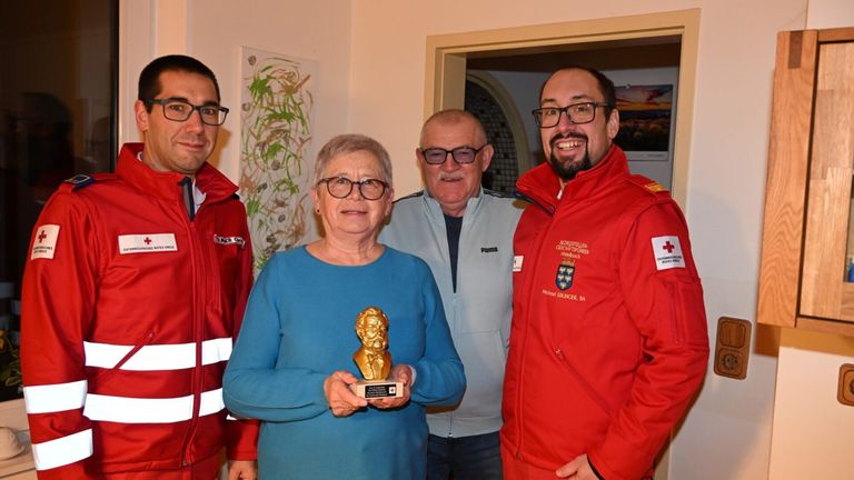 Eine Frau in einem blauen Shirt hält eine goldene Trophäe, umgeben von drei Männern in roten Jacken, die in der Nähe einer Wand mit Kunstwerken stehen.