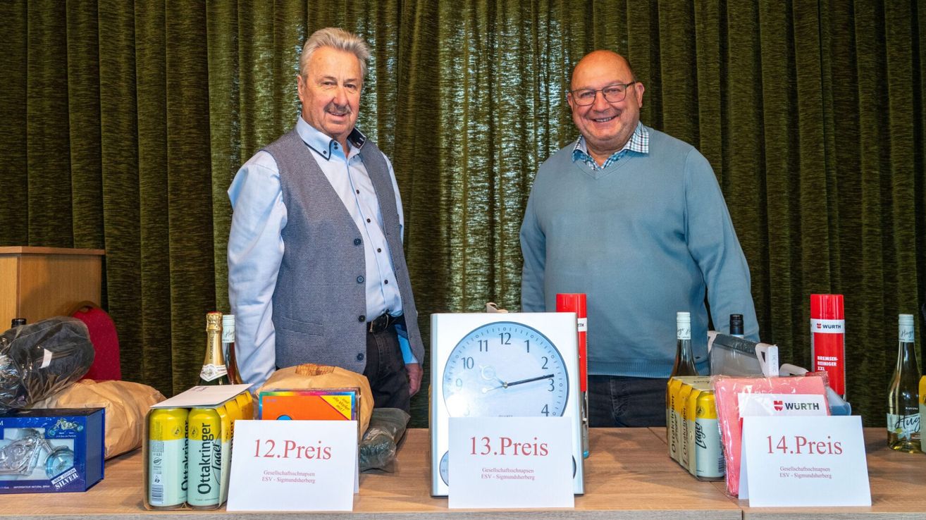 Zwei Männer stehen hinter einem Tisch mit Auktionsgegenständen, darunter eine Uhr, Weinflaschen und eine Schachtel. Der Mann links trägt eine Weste und der Mann rechts trägt eine Brille.